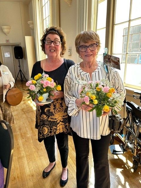 Two women holding flowers in front of a wooden floor.